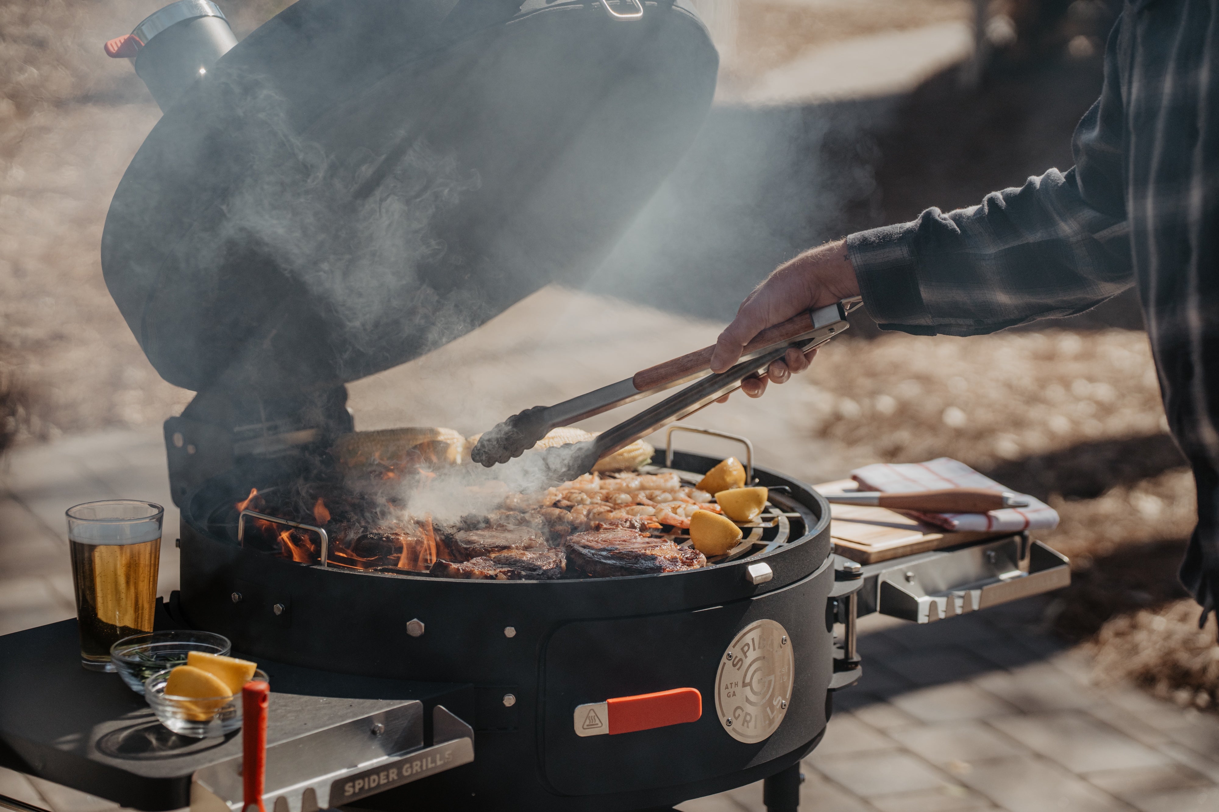 man cooking meat on open hunstman grill with beer and cutting boards on grill side tables