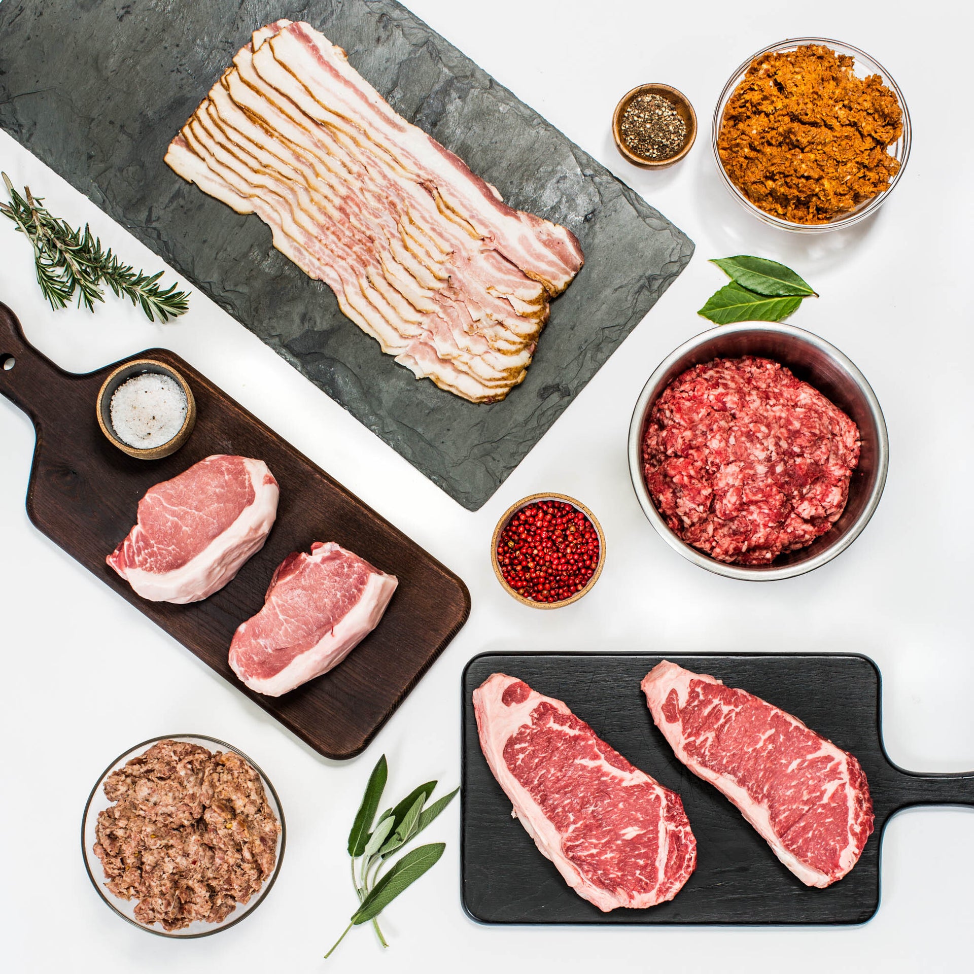 Assorted raw meats and seasonings including bacon, pork chops, ground beef, strip steaks, peppercorns, and fresh herbs